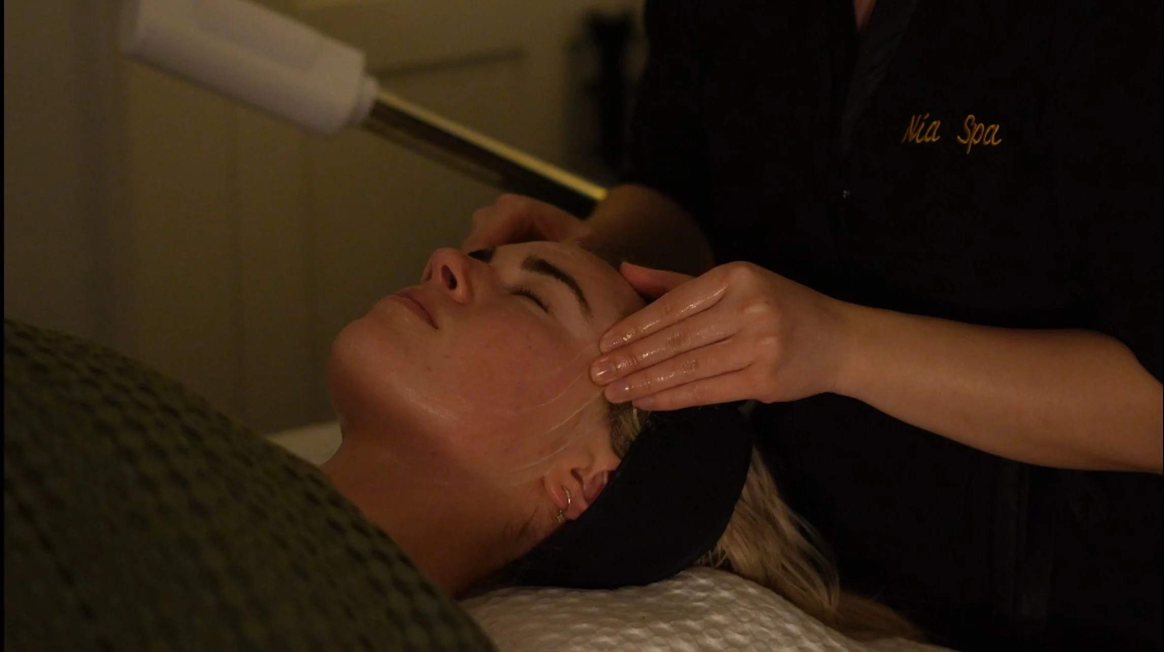 A woman receiving a facial massage at Nia Spa, lying on a bed with her eyes closed, while a therapist gently massages her face.