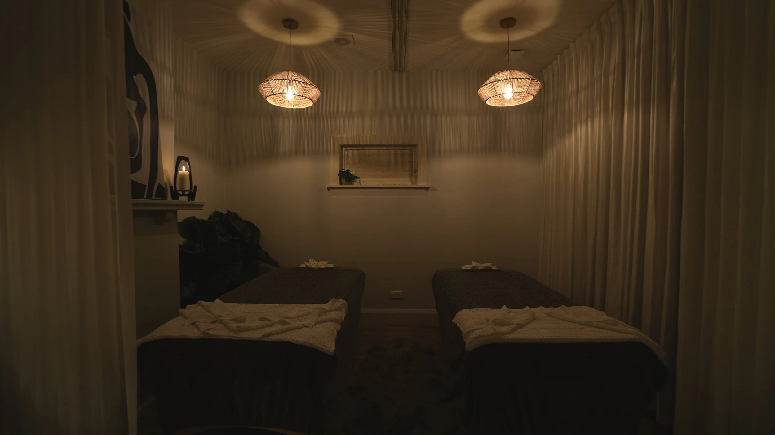 Two massage beds in a dimly lit spa room with pendant lights, curtains, and decorative wall art.