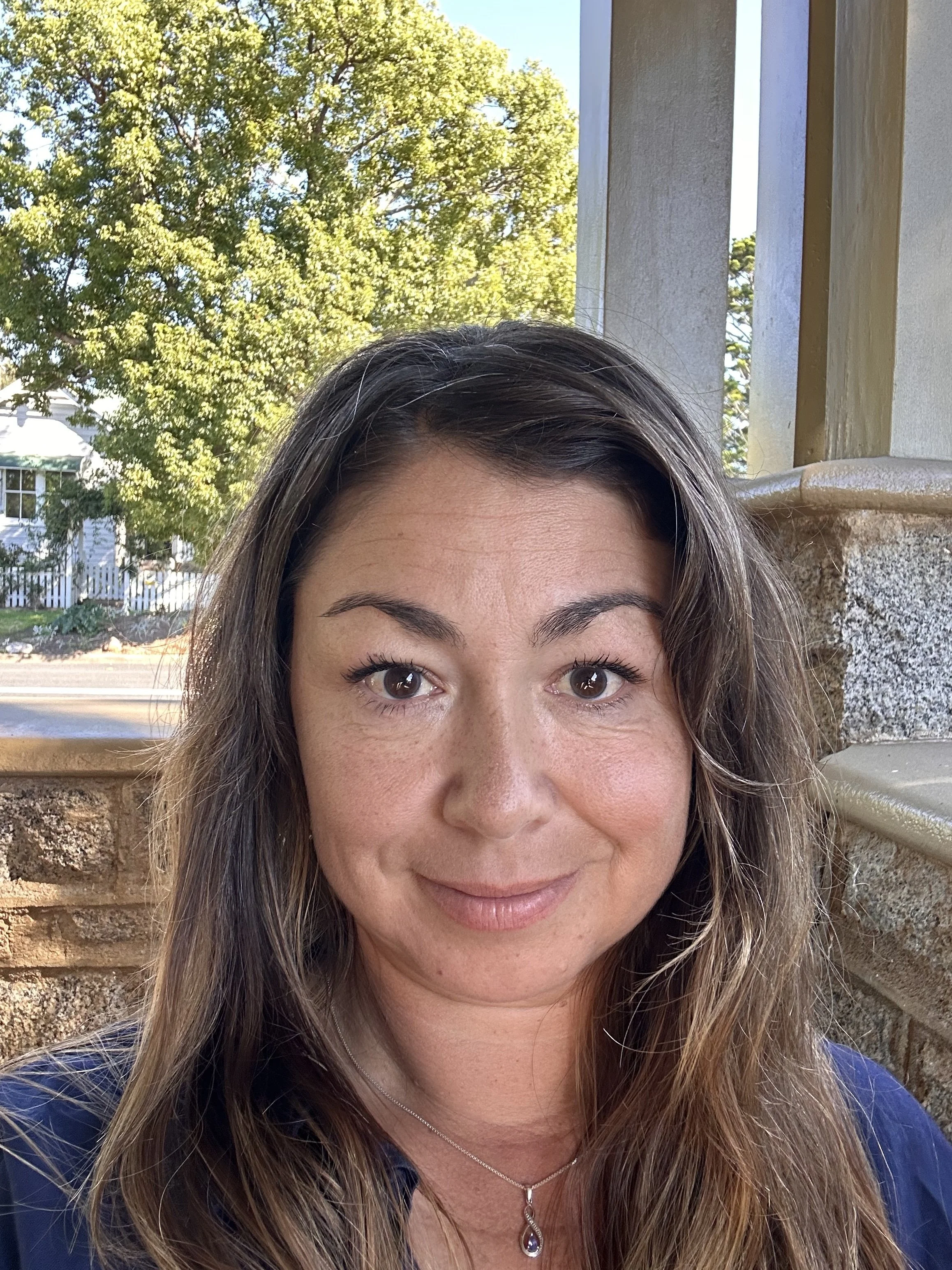 Close-up of a woman smiling outdoors, showing her face from the nose up, with brown eyes, dark eyebrows, and wavy brown hair with highlights. She is wearing a necklace with a purple gemstone pendant and a blue top. The background shows a sunny sky, green trees, and part of a house with a white fence.
