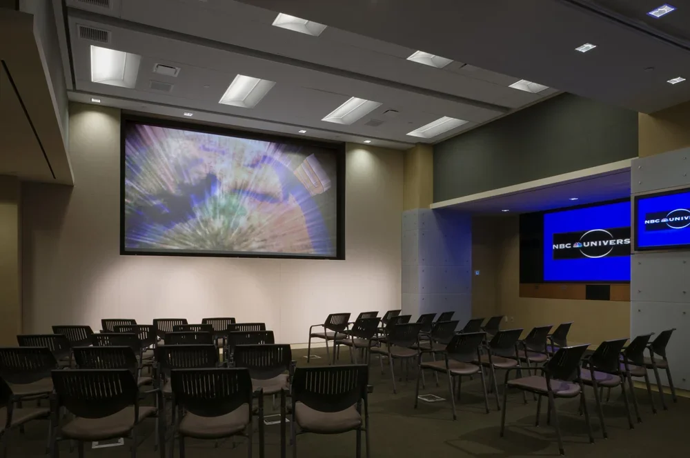 An empty conference room with chairs facing large screens displaying NBCUniversal logos, and a colorful abstract image on the main screen.