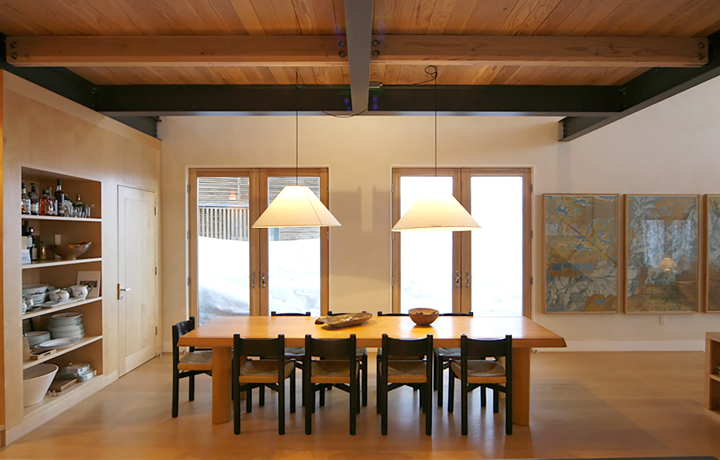 Dining room with a large wooden table, six chairs, three hanging pendant lights, and large glass doors leading outside, with a built-in shelf filled with dishes and bottles on the left, and three framed abstract paintings on the right wall.