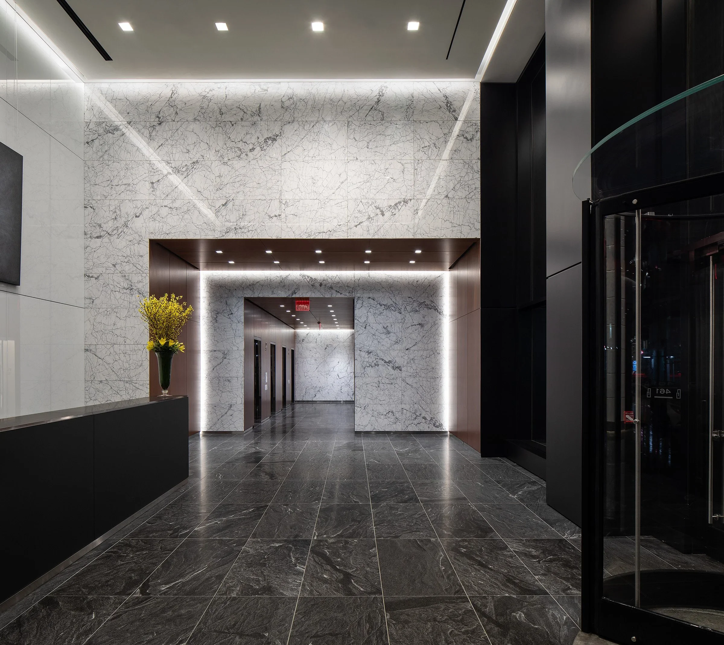 Modern hotel lobby with marble walls and floors, black accents, a vase with yellow flowers on a black counter, and a corridor leading to elevators and glass doors.