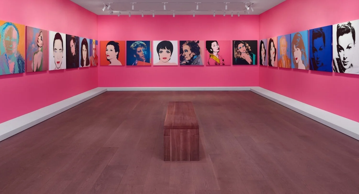 An art gallery with pink walls displaying colorful portraits of a woman, possibly Marilyn Monroe, on the walls, and a wooden bench in the center of the room.