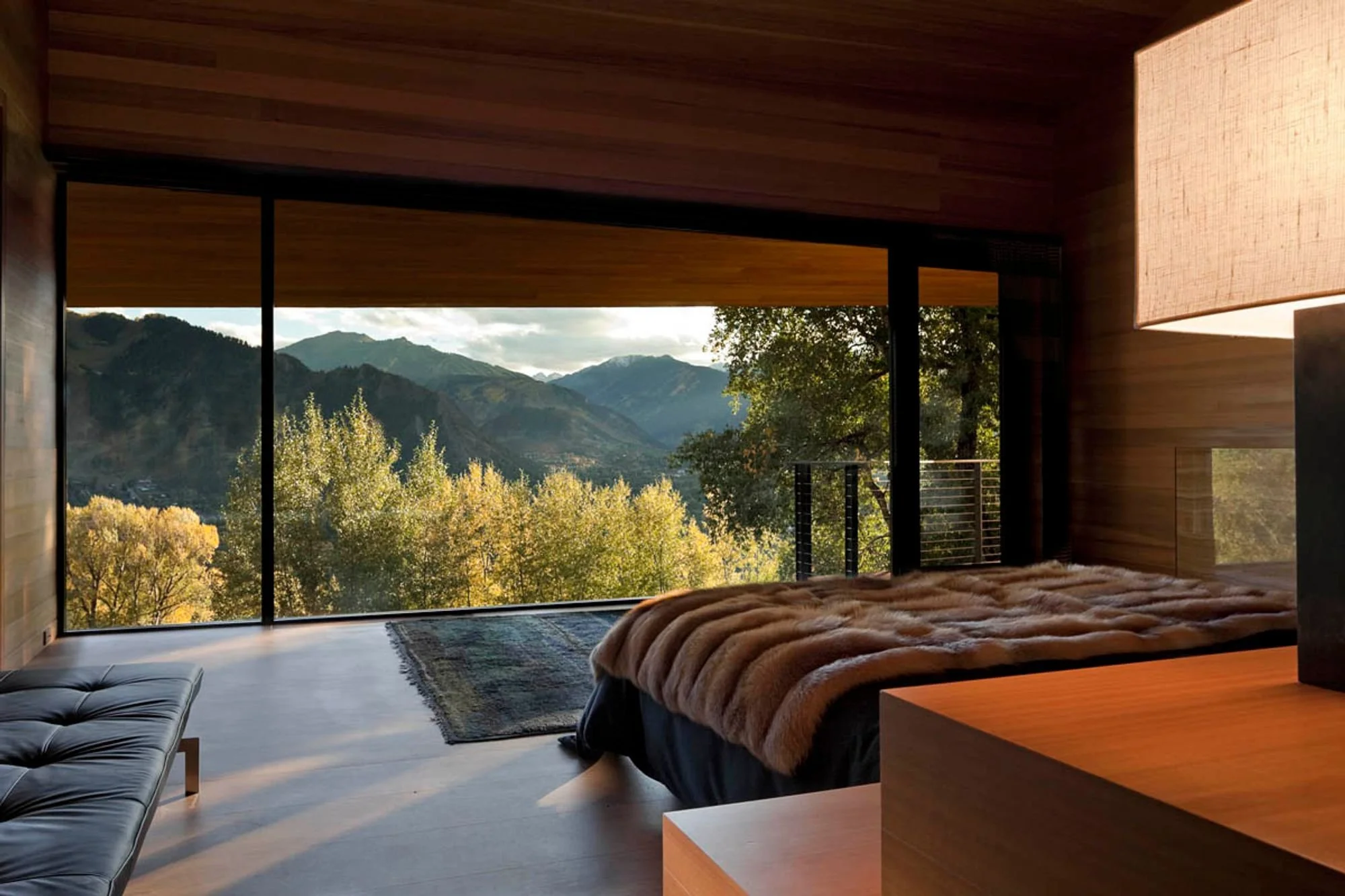 Modern hotel room with floor-to-ceiling windows revealing a scenic view of mountains and trees, a bed with a fur blanket, a wooden nightstand, and a lamp.
