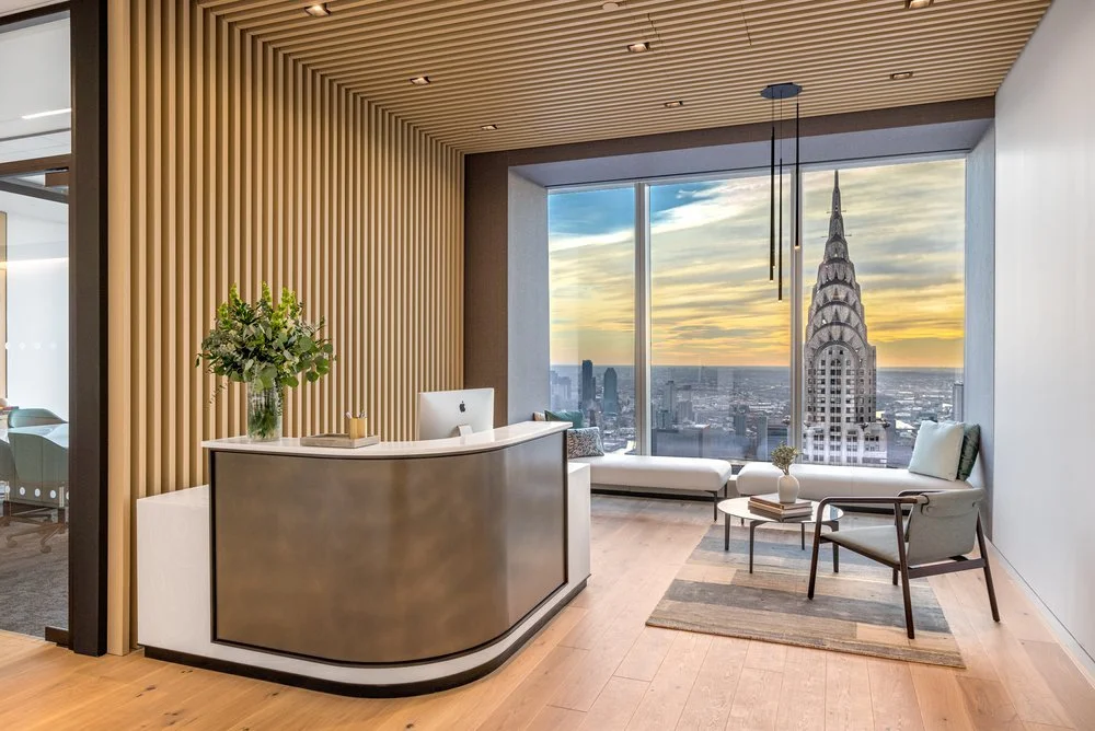 Modern office with a curved reception desk, large window view of the New York City skyline and the Chrysler Building at sunset, seating area with chairs, a small table, and decorative plants.