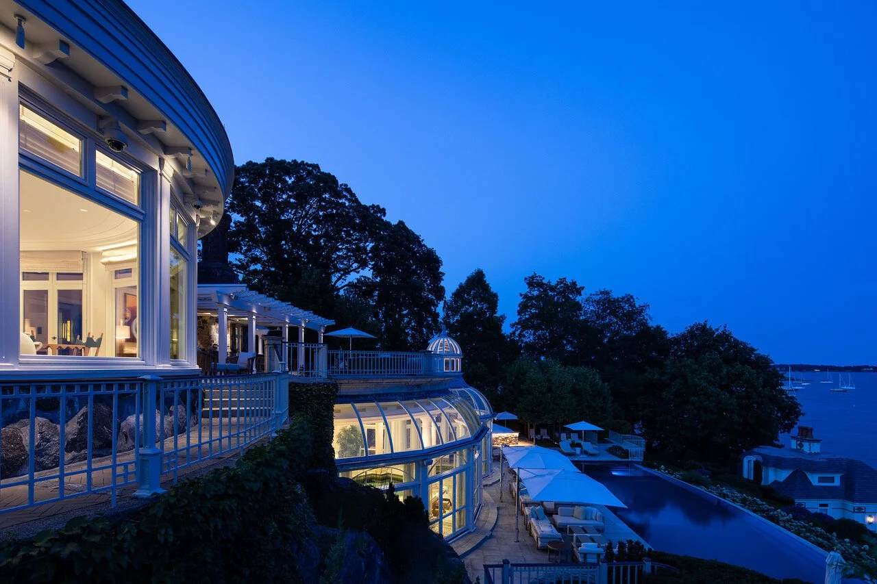 A luxurious house with large glass windows, illuminated from inside, featuring outdoor seating, a pool, and a view of the water with boats, during dusk.