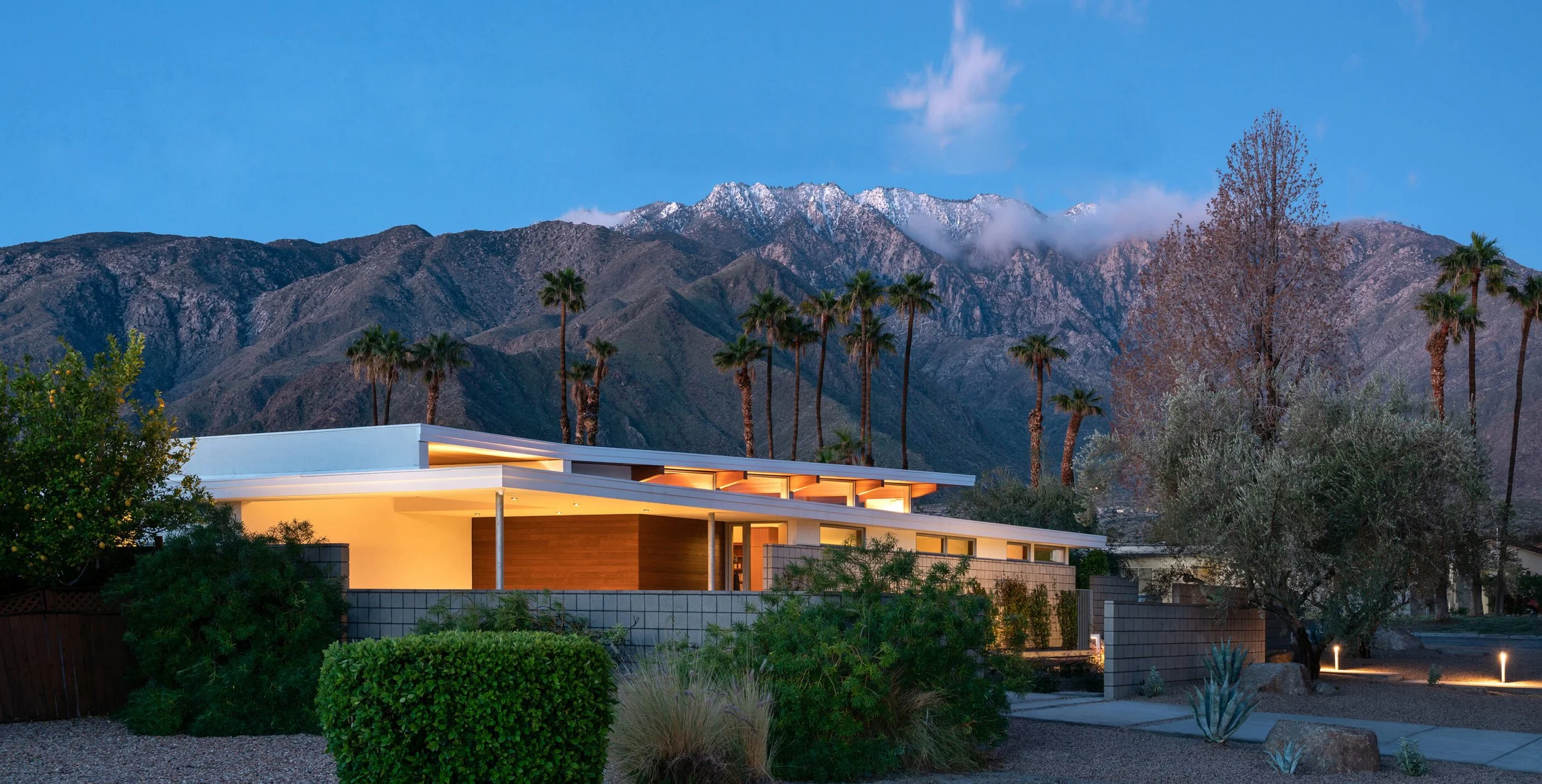 Modern house in a desert landscape with mountains and palm trees in the background, illuminated at dusk.