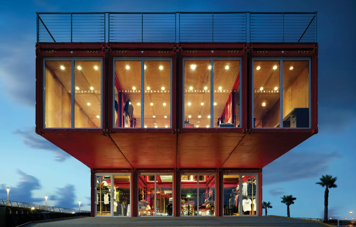 A modern, stacked container building with large glass windows, illuminated from inside, houses retail spaces with clothing displays and interior lighting at dusk. Palm trees line the background.