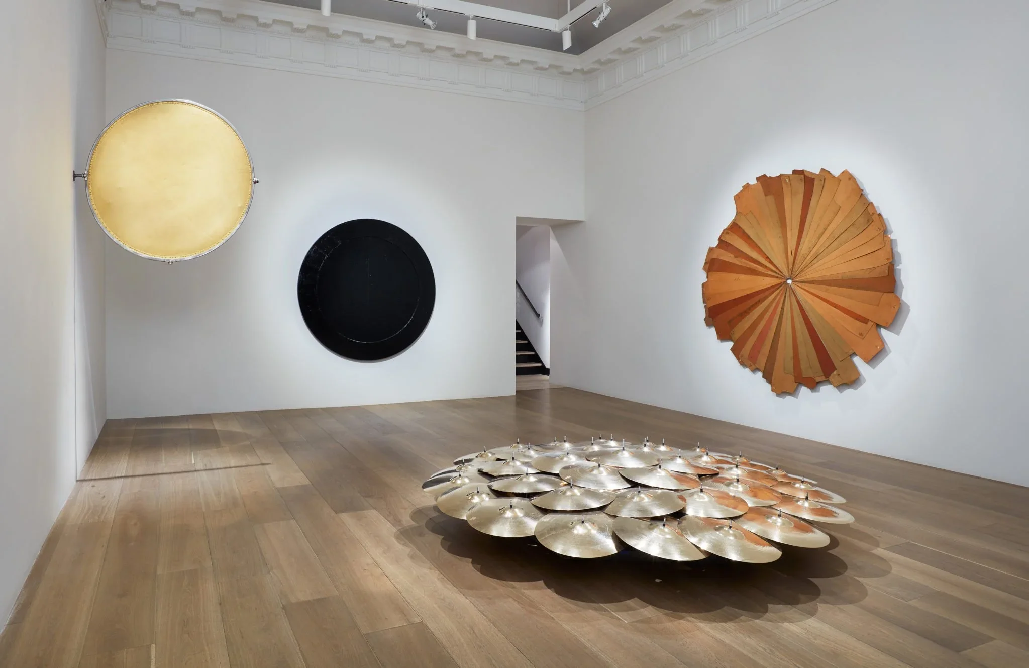 Contemporary art installation in a gallery featuring large circular artworks on a white wall and a sculptural piece made of semi-circular metallic plates on the wooden floor.