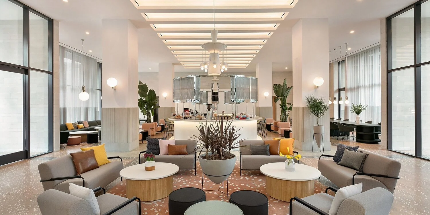 A spacious hotel lobby with modern decor, large windows, and comfortable seating arrangements including sofas, armchairs, and potted plants. There is a central bar with glass partitions and a high ceiling with recessed lighting.