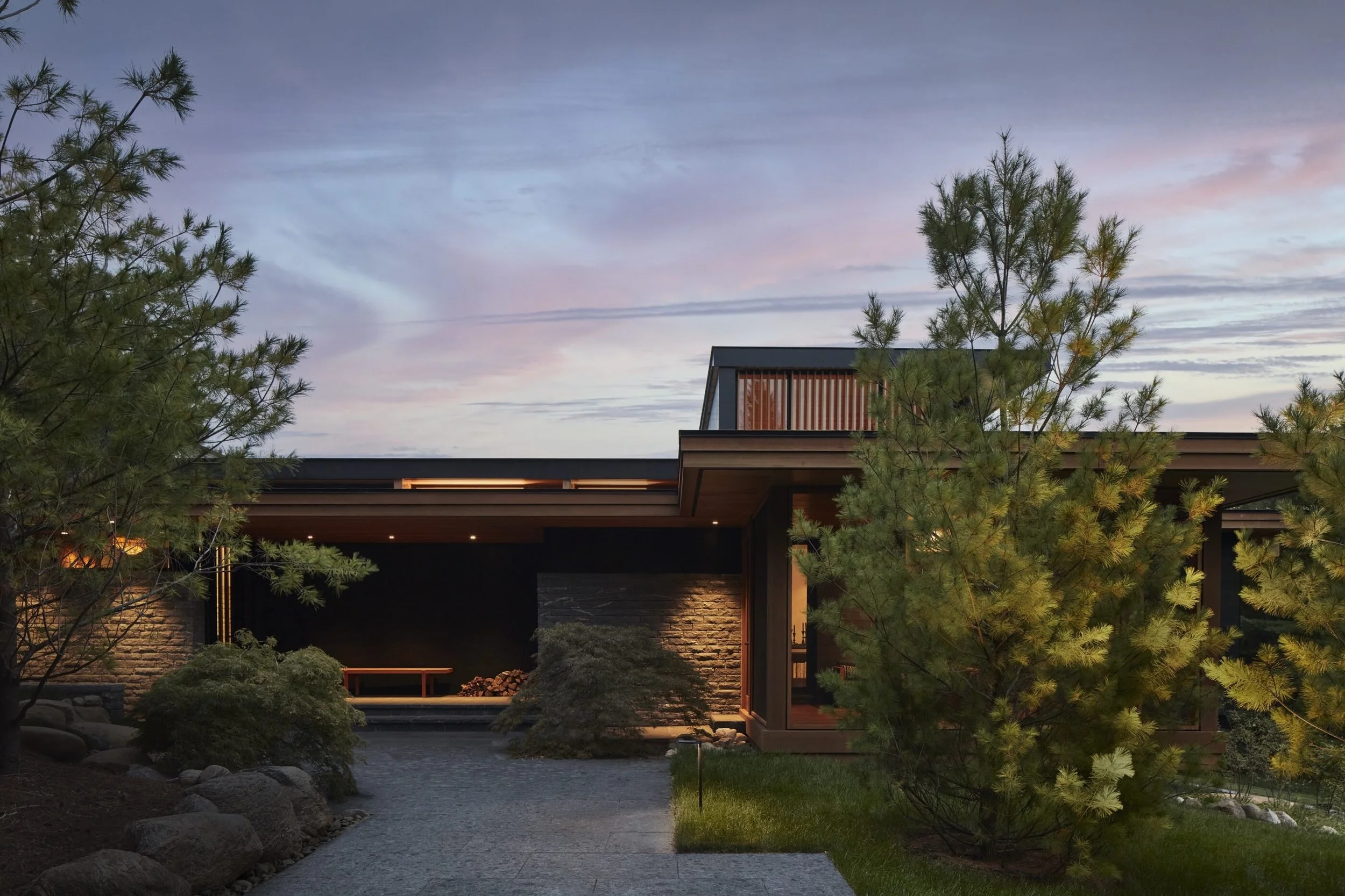 Modern house with large windows, surrounded by trees and landscaping, during dusk