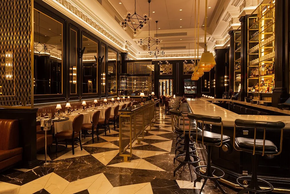 Elegant bar and dining area with black and gold accents, marble floor, high ceilings, and modern light fixtures.