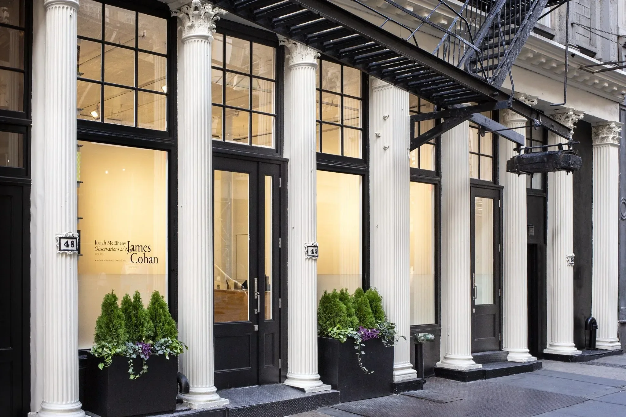 Exterior view of an art gallery with classical columns, large window panes, black doors, and decorative metal fire escape stairs, with potted plants outside the entrance.