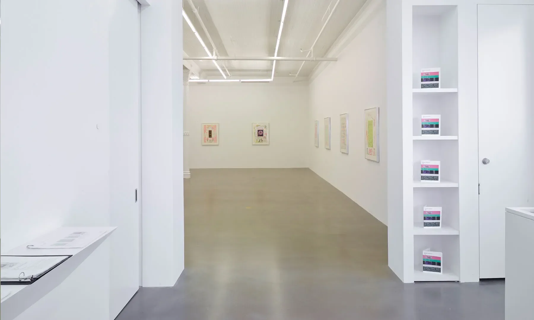 View of an art gallery with white walls, framed artwork, and a counter with brochures or catalogues on the left side.