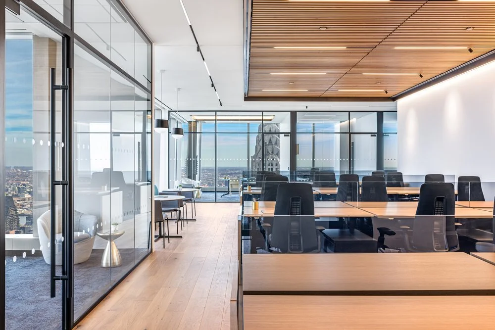 Modern office conference room with large glass windows, wooden ceiling, and multiple desks with black chairs, overlooking a cityscape.