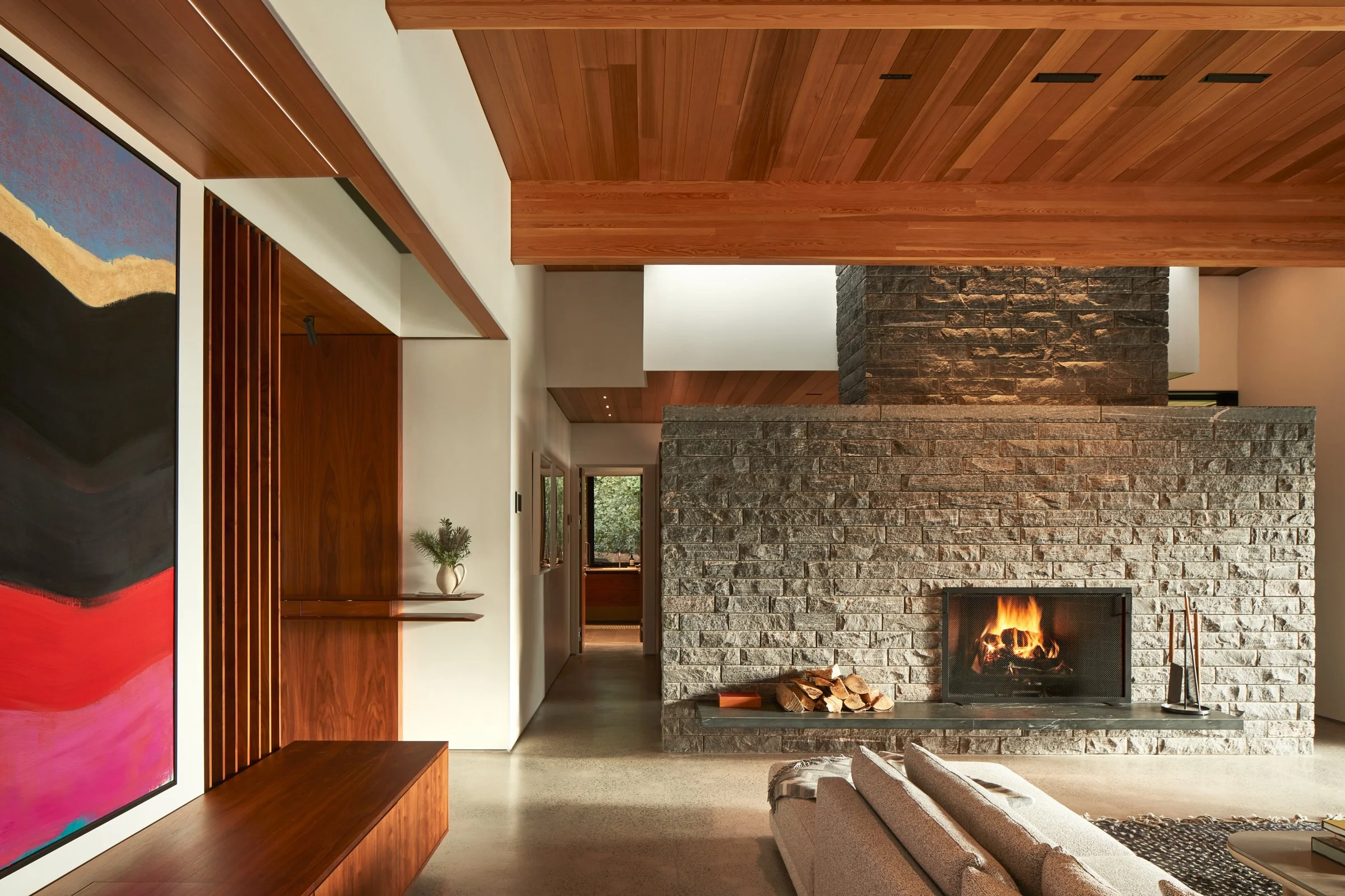 Modern living room with a large stone fireplace, wood-paneled ceiling, abstract colorful artwork on the wall, and a beige sofa.