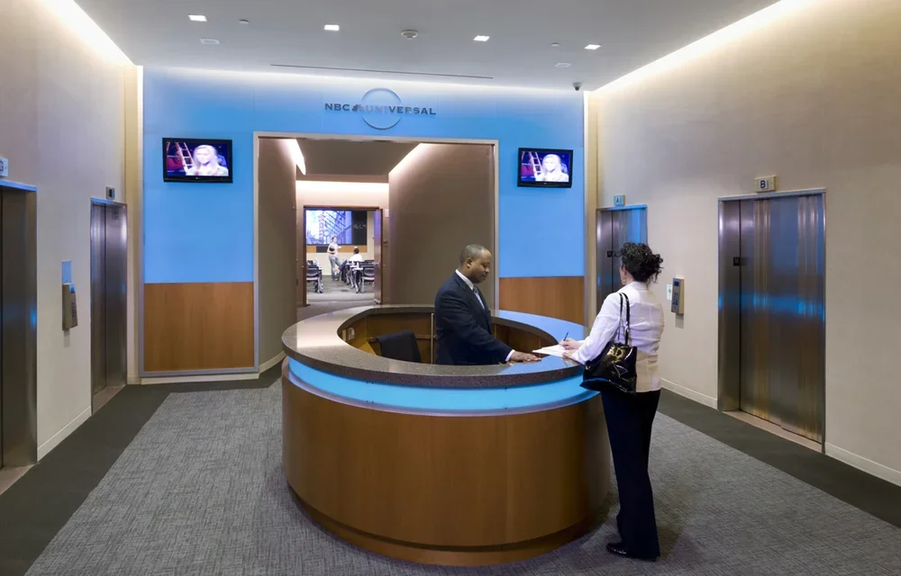 Reception area at NBCUniversal with a circular desk where a man is assisting a woman, two elevators on either side, and two TV screens showing a woman speaking.