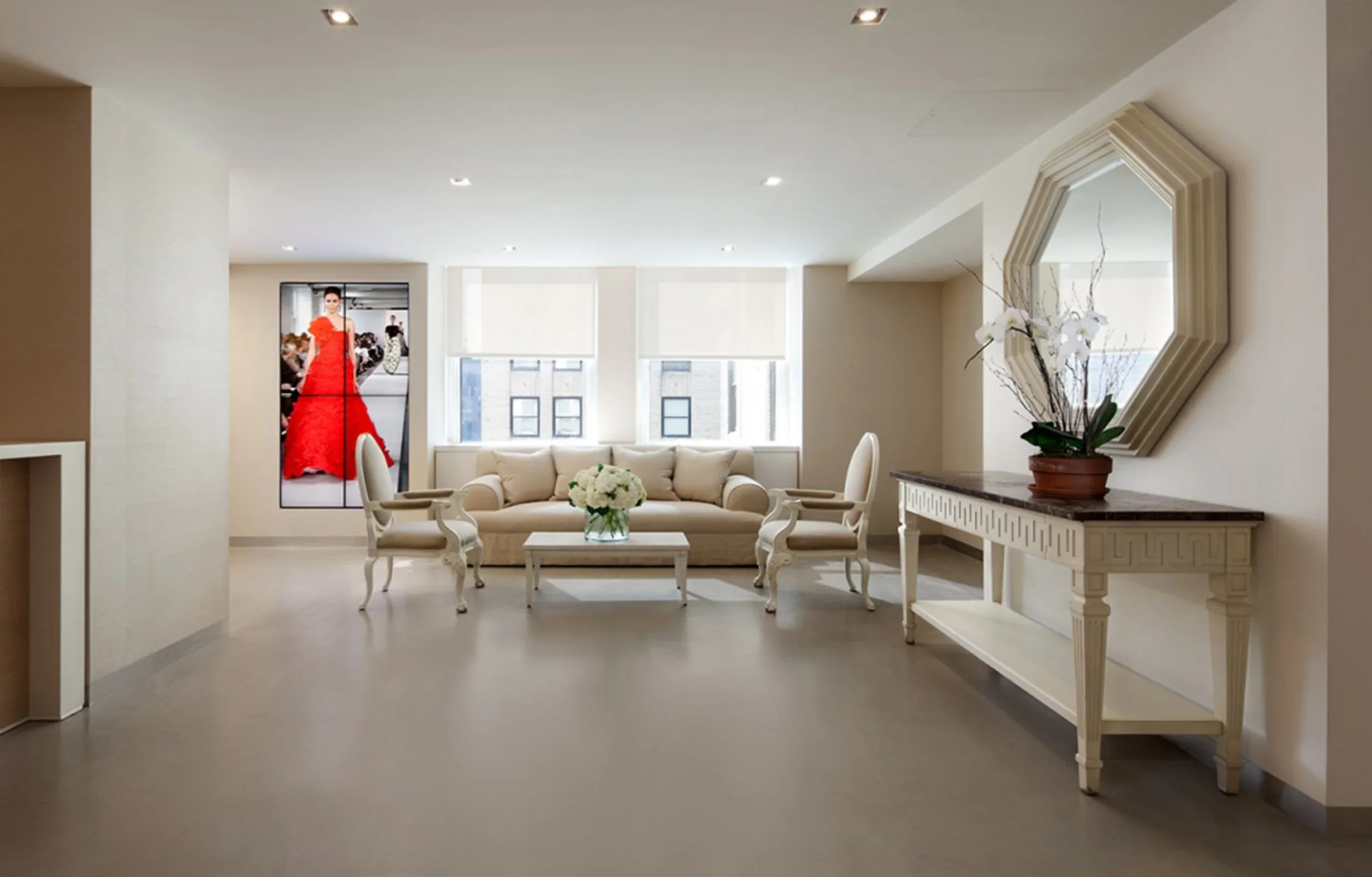 Modern minimalist living room with beige sofa, two armchairs, a white coffee table with flower arrangement, decorative mirror, flowering plant, and a digital photo frame on the wall, bright with three large windows.