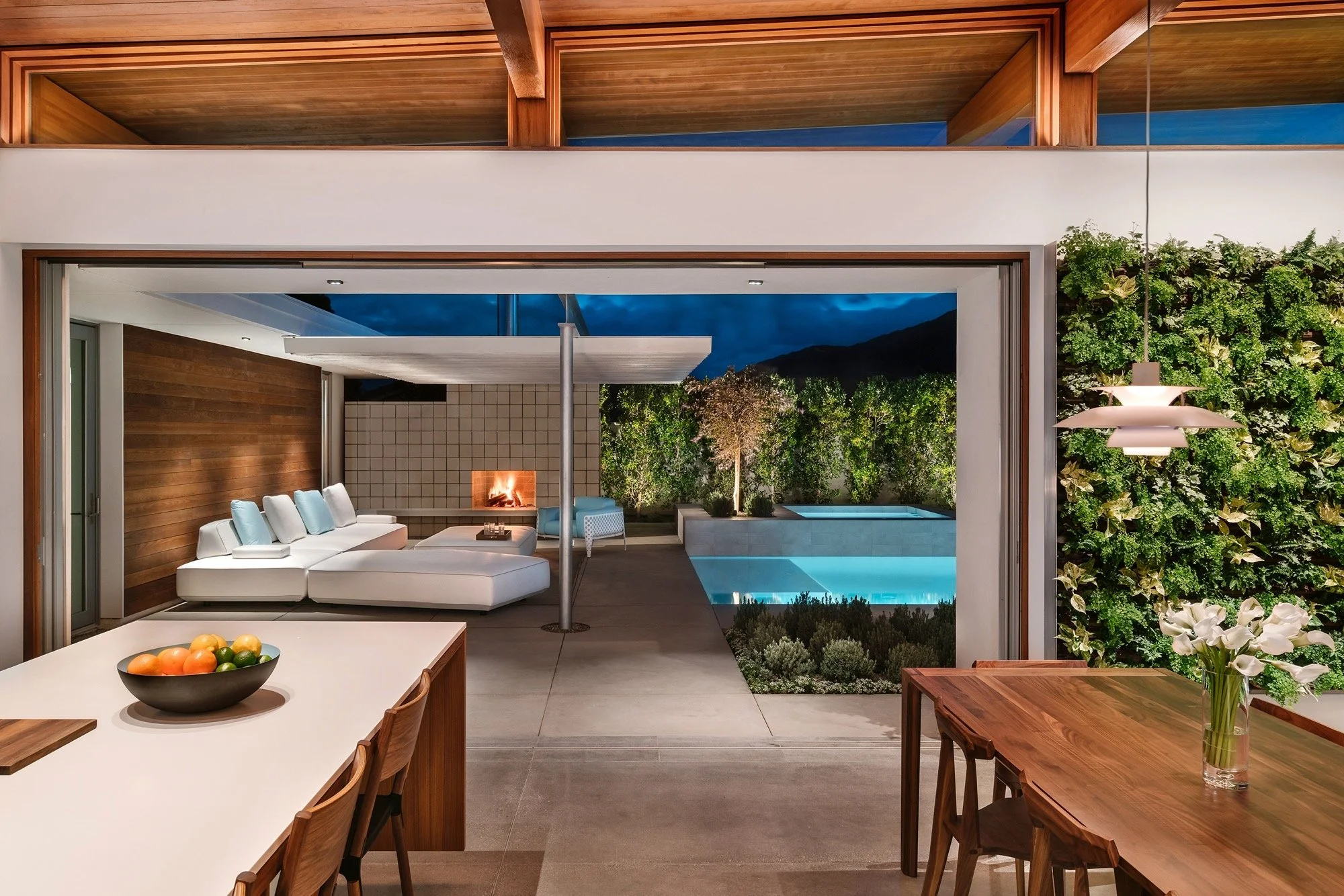 Modern open-plan living area with wooden accents, a white sectional sofa with light blue pillows, a fireplace, an outdoor pool, and greenery at night.