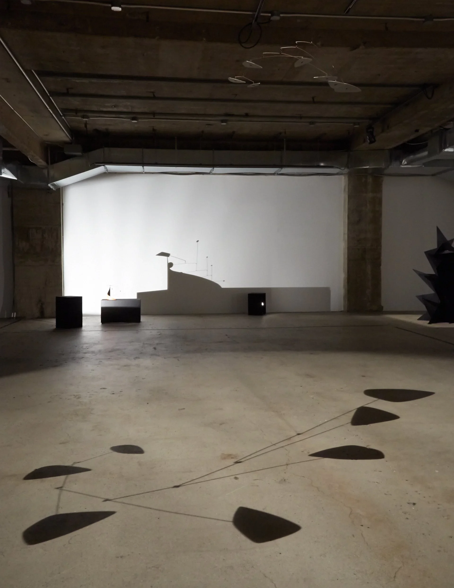 An art installation in a spacious industrial-style gallery features a large white screen displaying shadows cast by various abstract sculptures and objects, with additional sculptures and shadows visible on the floor and walls.