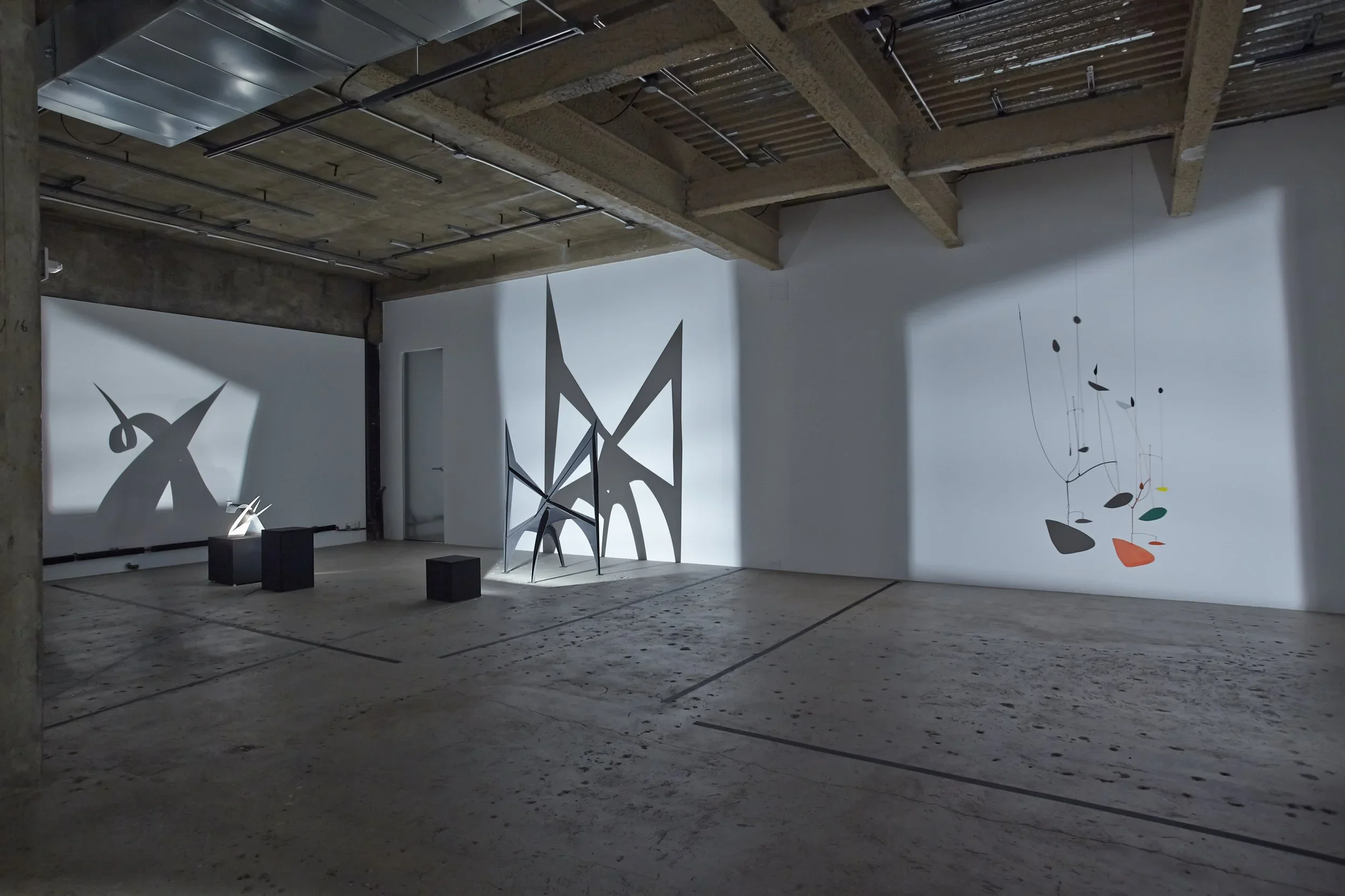 Art installation with abstract sculptures casting shadows and a mobile hanging with colorful shapes in an industrial gallery space.