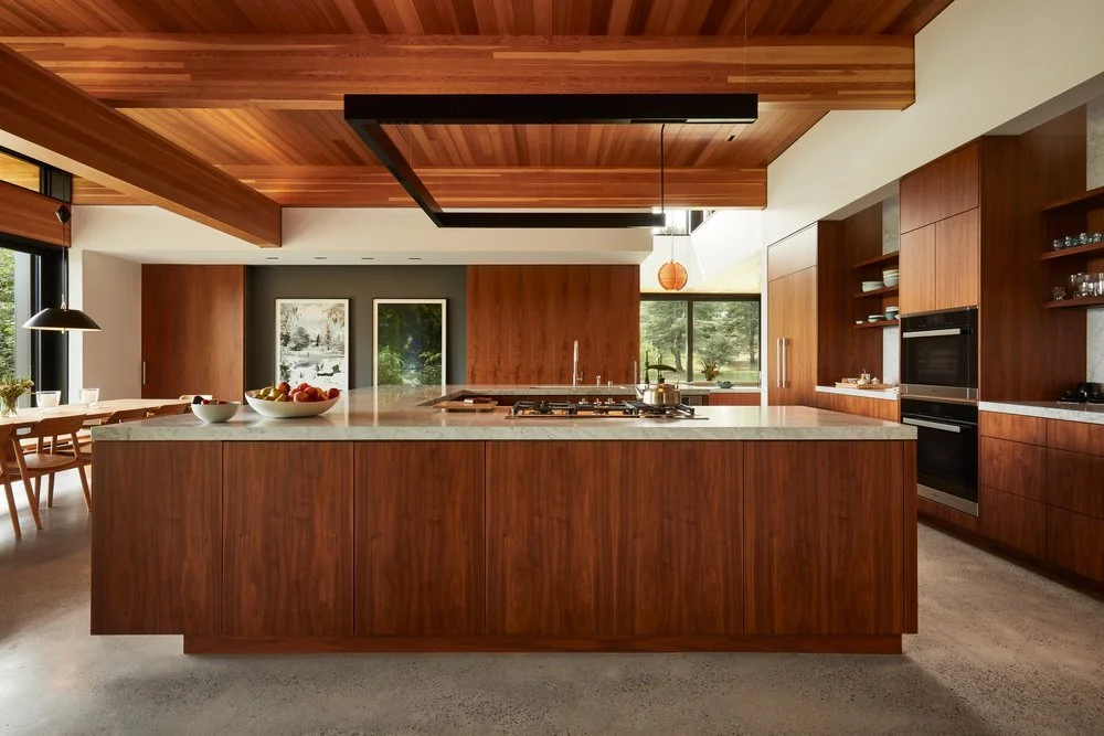 Modern kitchen with wooden cabinets, white marble island, and built-in oven. Large windows with views of outdoors, pendant lighting, and dining area with chairs and table.