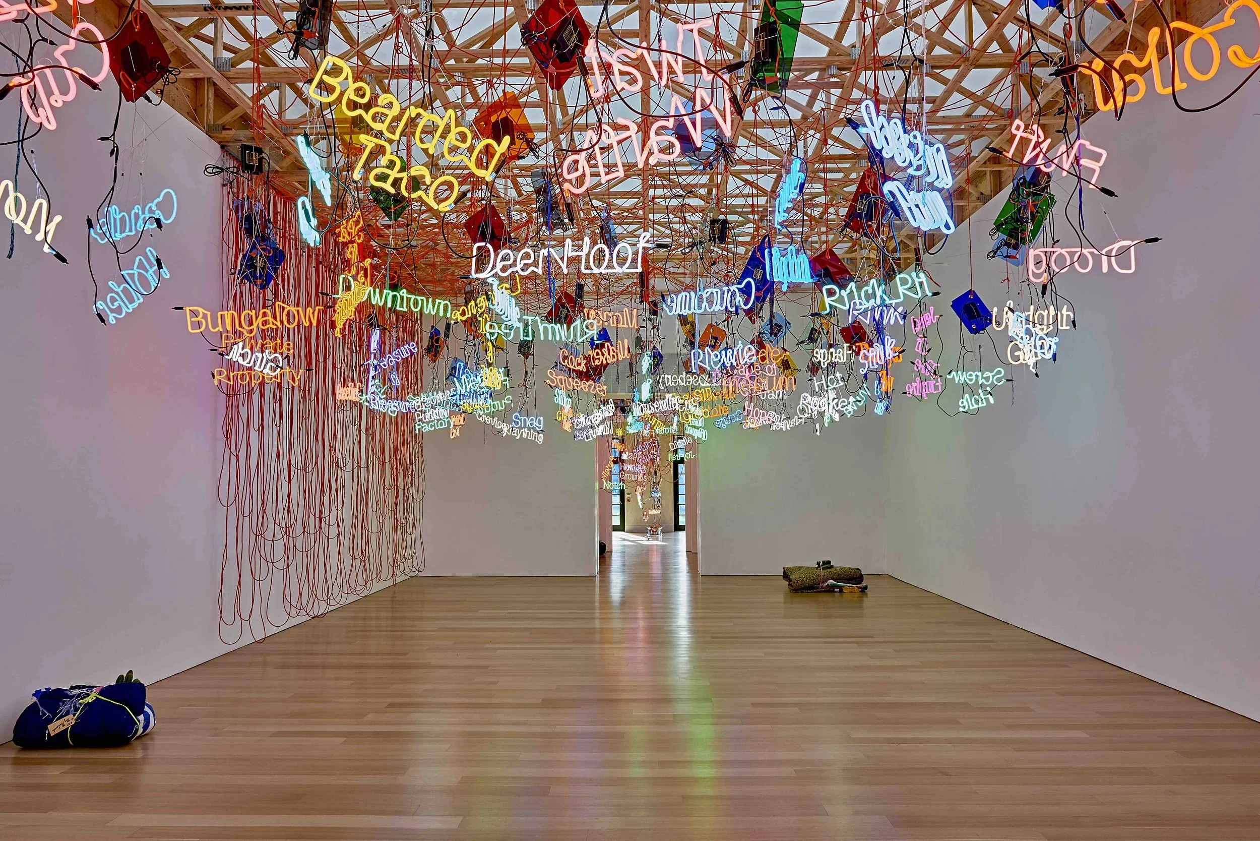 An art installation featuring colorful neon signs hanging from a wooden ceiling framework inside a gallery space with wooden floor. The signs display various words and phrases in different colors and styles.