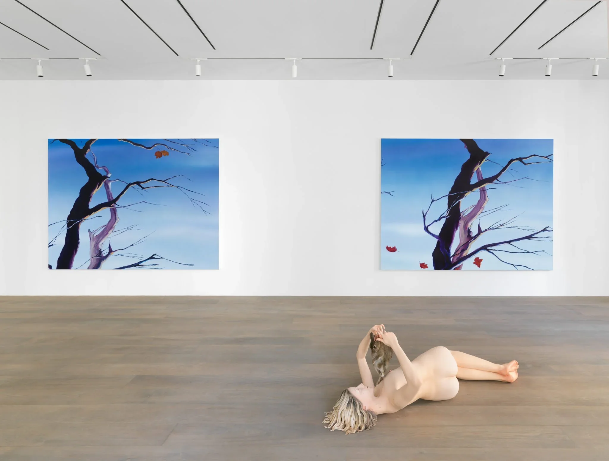 A nude woman lying on her back on a wooden floor in an art gallery, holding a small animal above her face. Two paintings of leafless trees against a blue sky with red leaves are displayed on the white wall behind her.