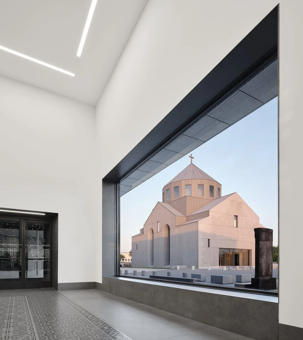 The photo shows the interior of a modern building with white walls, a large window, and a traditional church with a dome and cross outside.