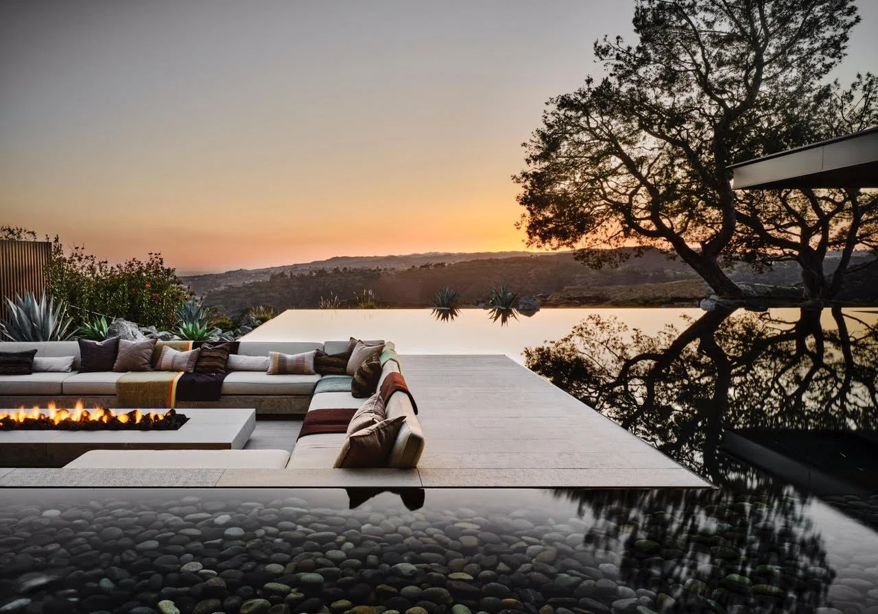 Modern outdoor patio with a firepit surrounded by cushioned seating, overlooking a landscape at sunset with a tree and an infinity pool reflecting the sky.
