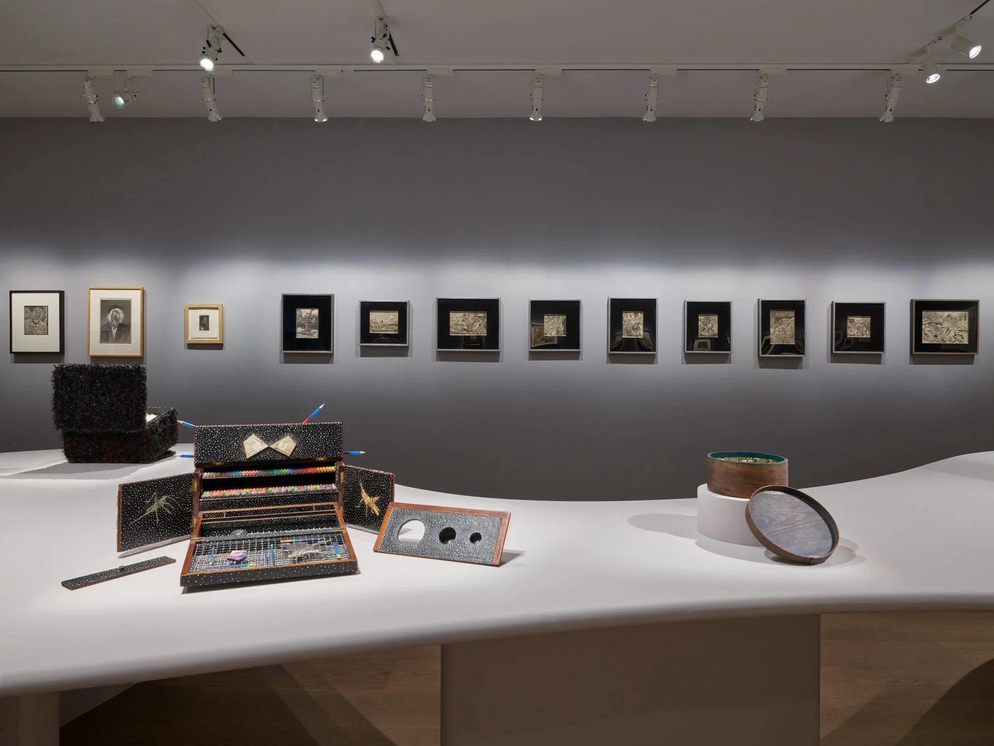Art gallery display featuring framed monochrome artworks on a wall, with jewelry and decorative objects on a white table in the foreground.