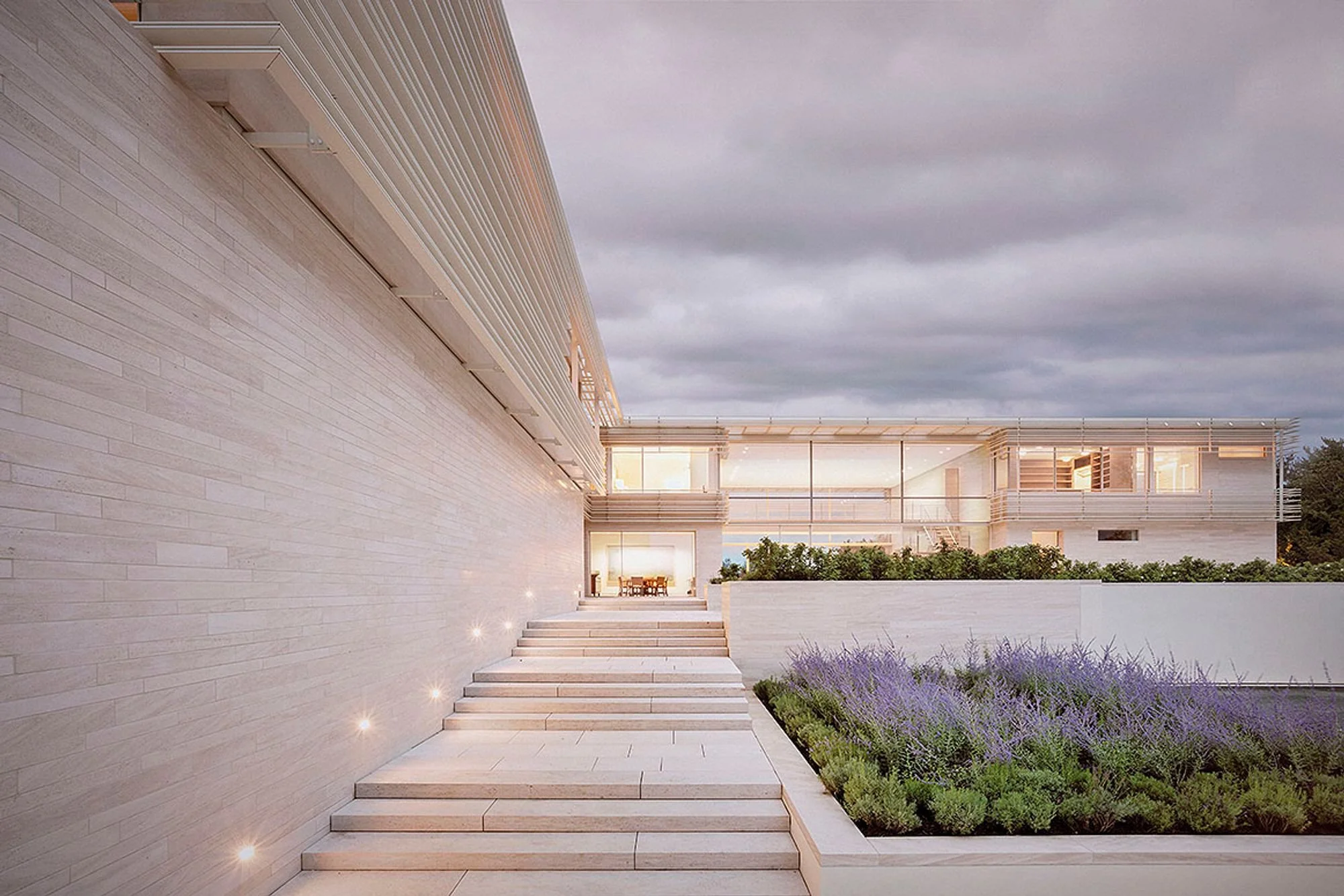 Modern building with large glass windows, outdoor seating, and a landscaped garden with purple flowers under a cloudy sky.