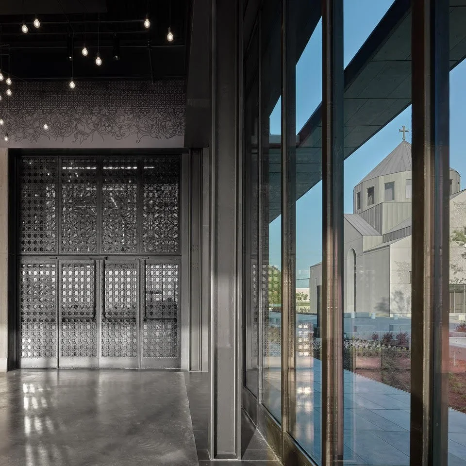 Interior view of a modern building with large glass windows and a view of a church with a cross on its roof outside.