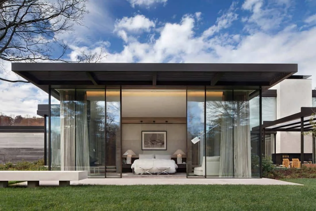 Modern house with glass walls, white curtains, and a bedroom visible through the glass, under a partly cloudy sky.