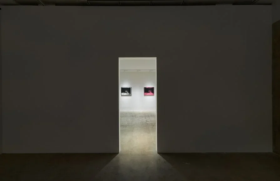 Museum or gallery corridor with two framed photographs on white wall, viewed through a rectangular doorway.
