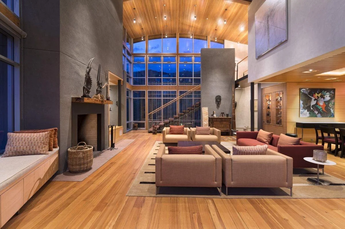 Spacious modern living room with a high wooden ceiling and large windows showing an evening sky. The room features a fireplace, contemporary sculptures, and seating with pillows, with a staircase leading upstairs.