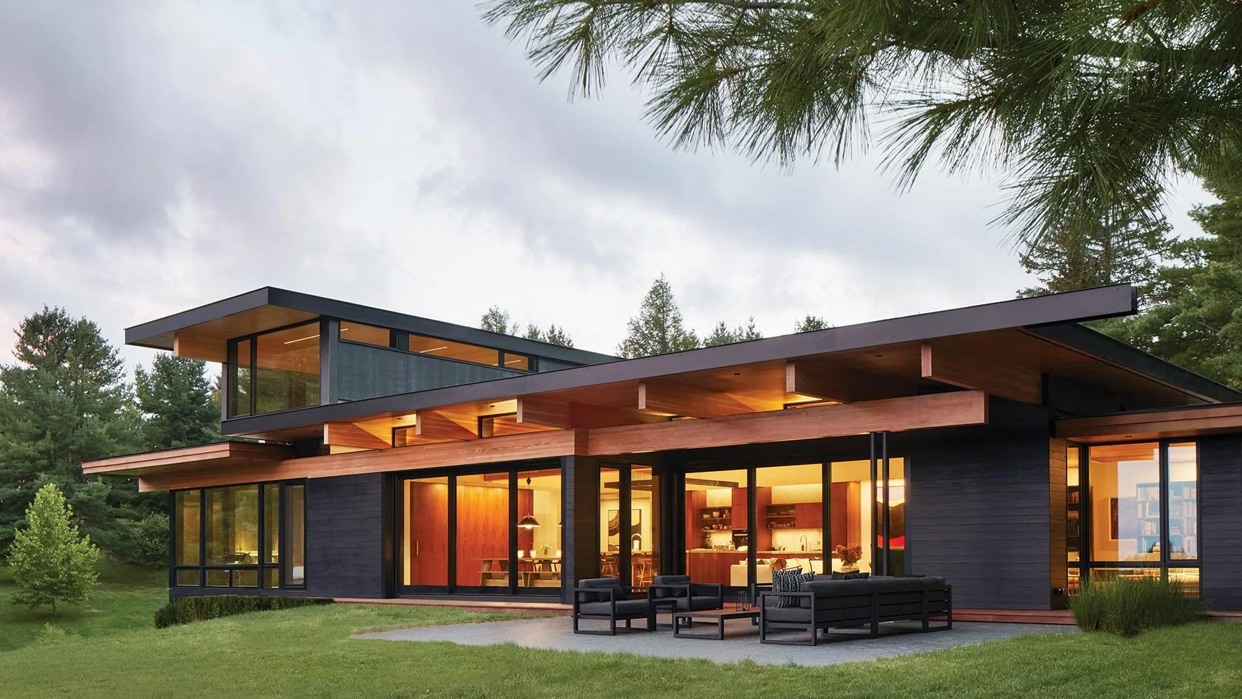 Modern house with large glass windows, black exterior, wooden accents, outdoor seating area, surrounded by grass and trees, evening lighting
