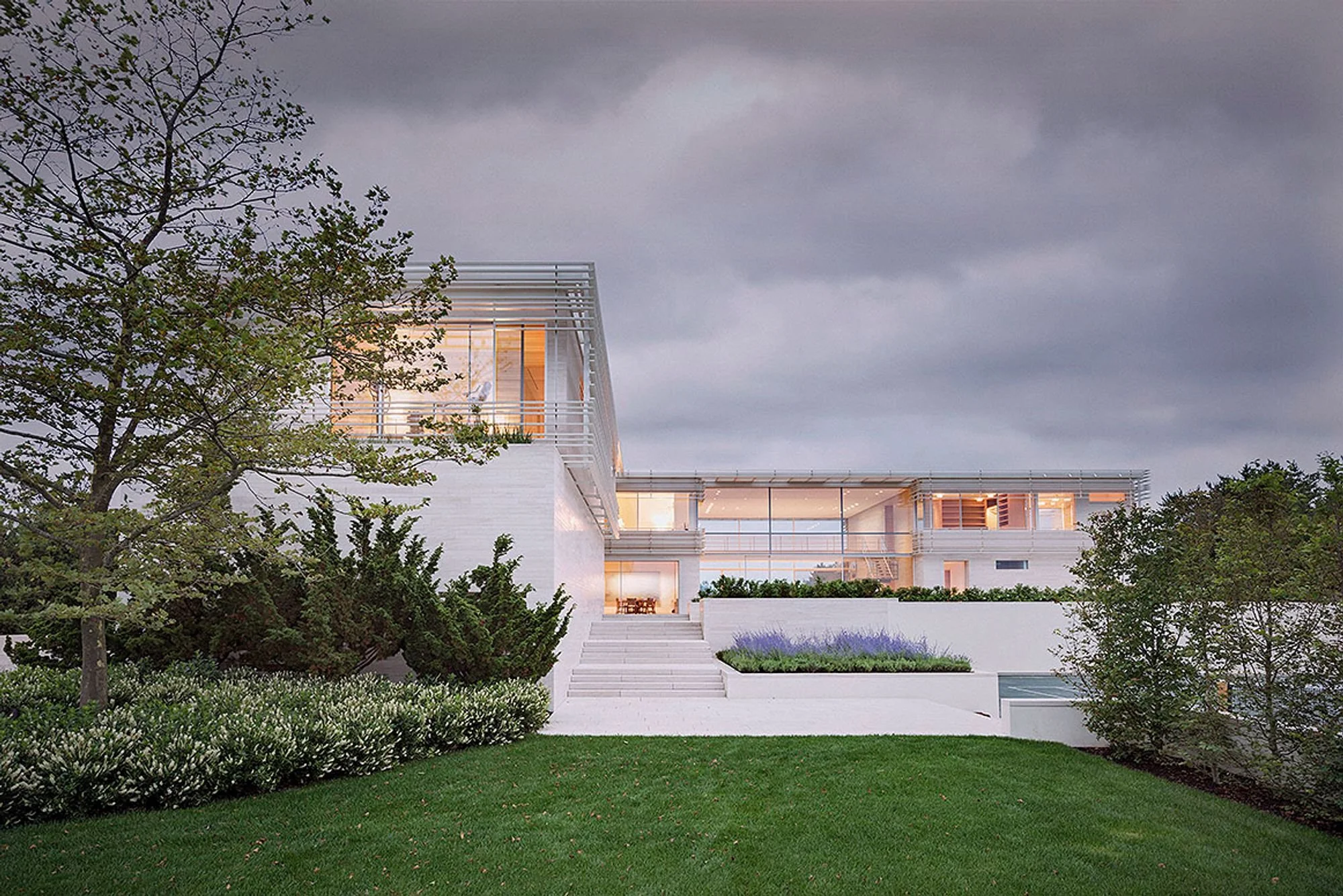 Modern white house with multiple levels, glass windows, and outdoor lighting, surrounded by landscaped greenery and trees under a cloudy sky.