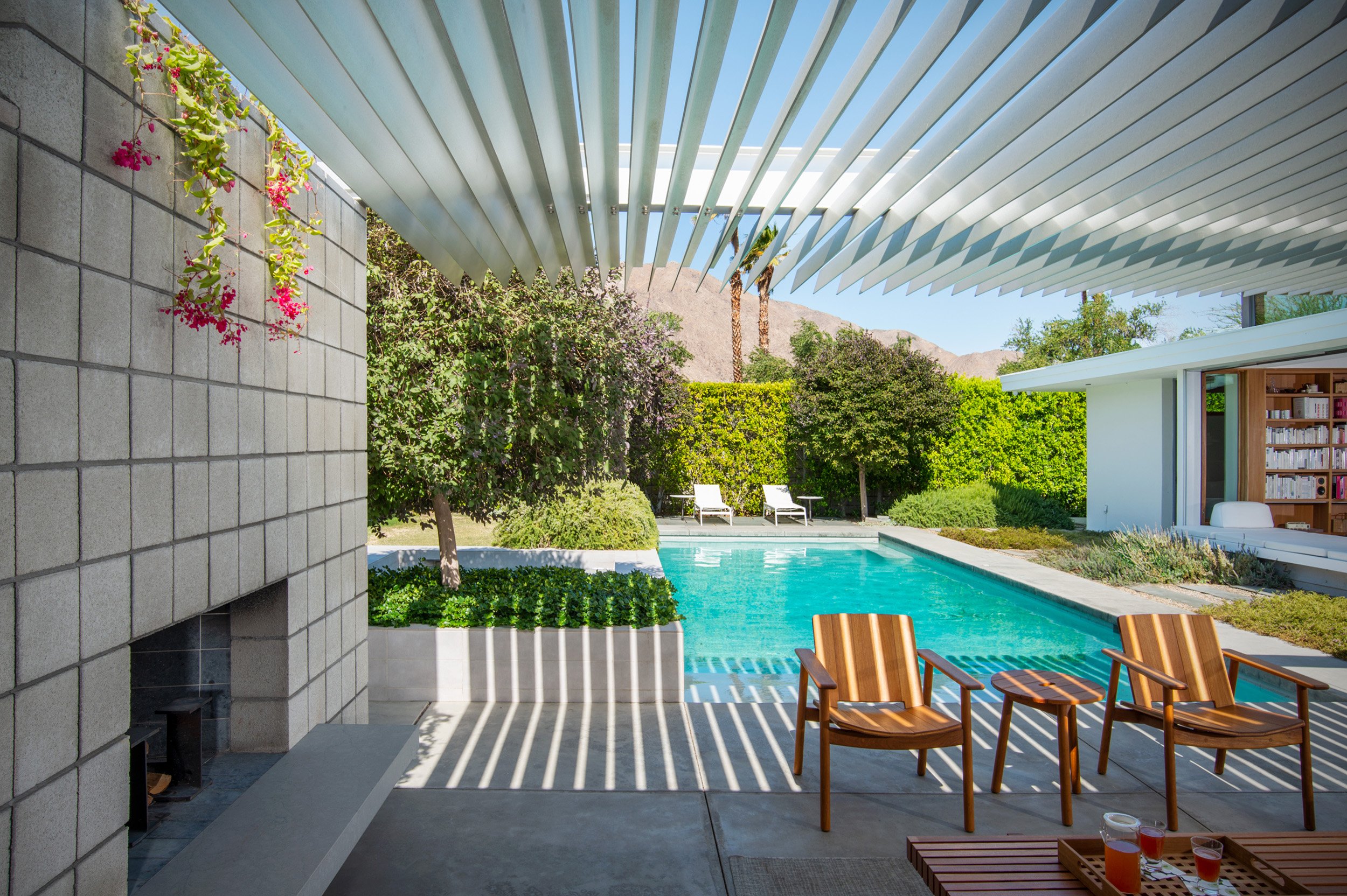 Modern backyard patio with a swimming pool, wooden chairs, lush green trees, and a house with bookshelves visible through an open sliding door. Sunlight creates striped shadows from a pergola overhead.