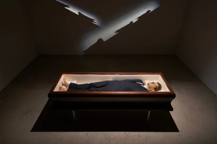 A wax figure of a man in a suit lying in a wooden coffin in a dimly lit room, with shadowy reflections cast on the wall above.