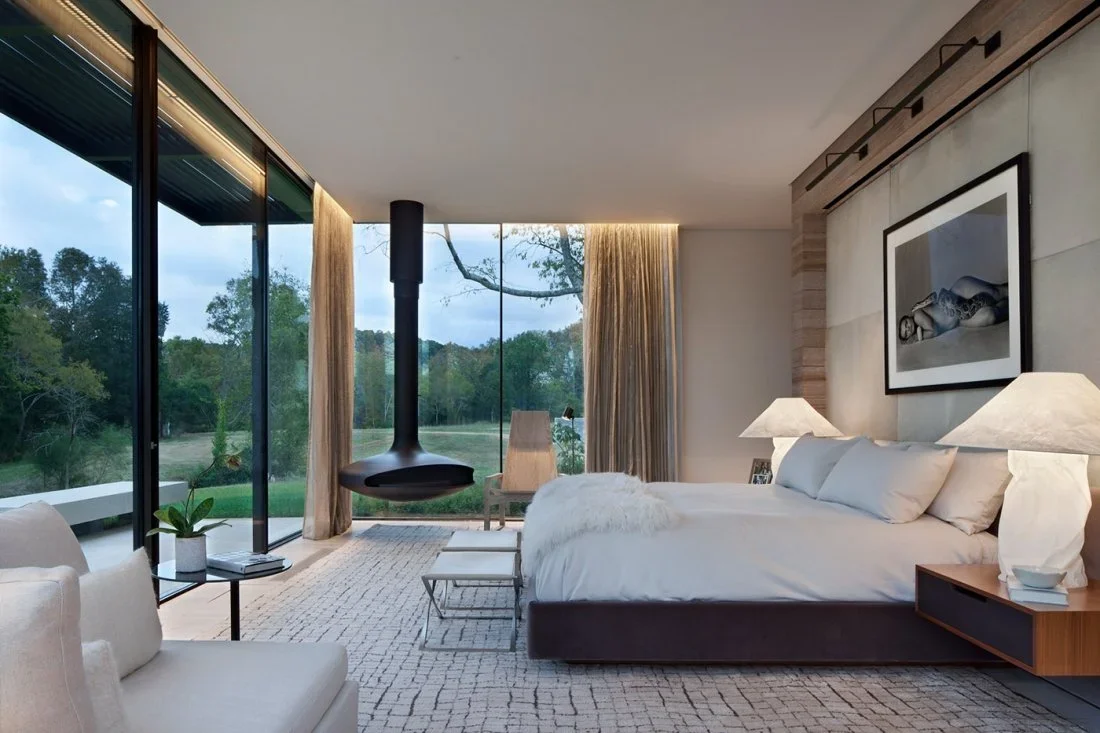Modern bedroom with large glass windows overlooking a green outdoor landscape, featuring a suspended fireplace, white bed with multiple pillows and lamps, a beige armchair, and a throw blanket.