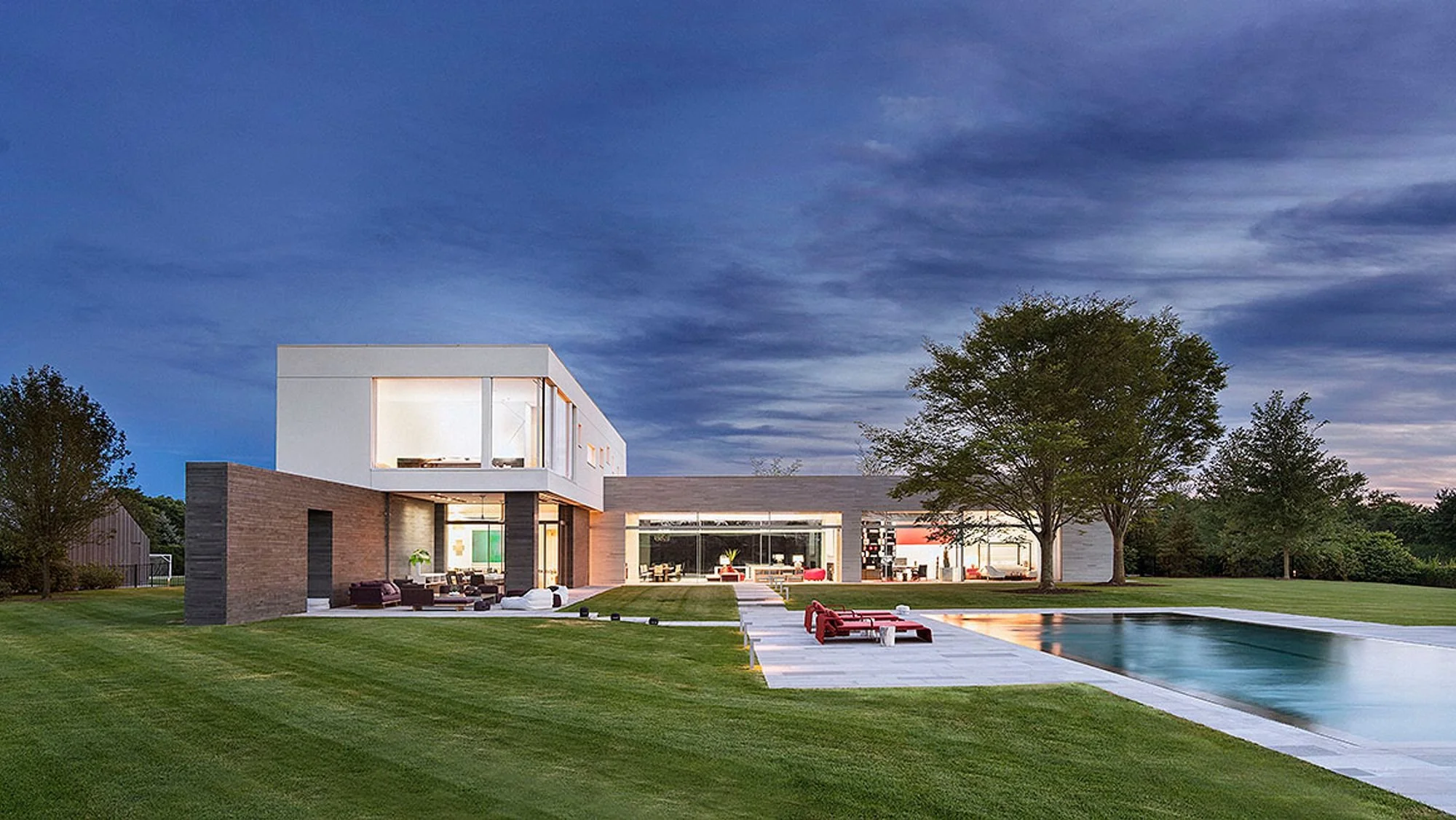 Modern house with large glass windows, outdoor seating area, swimming pool, and well-maintained lawn under a partly cloudy sky at dusk.