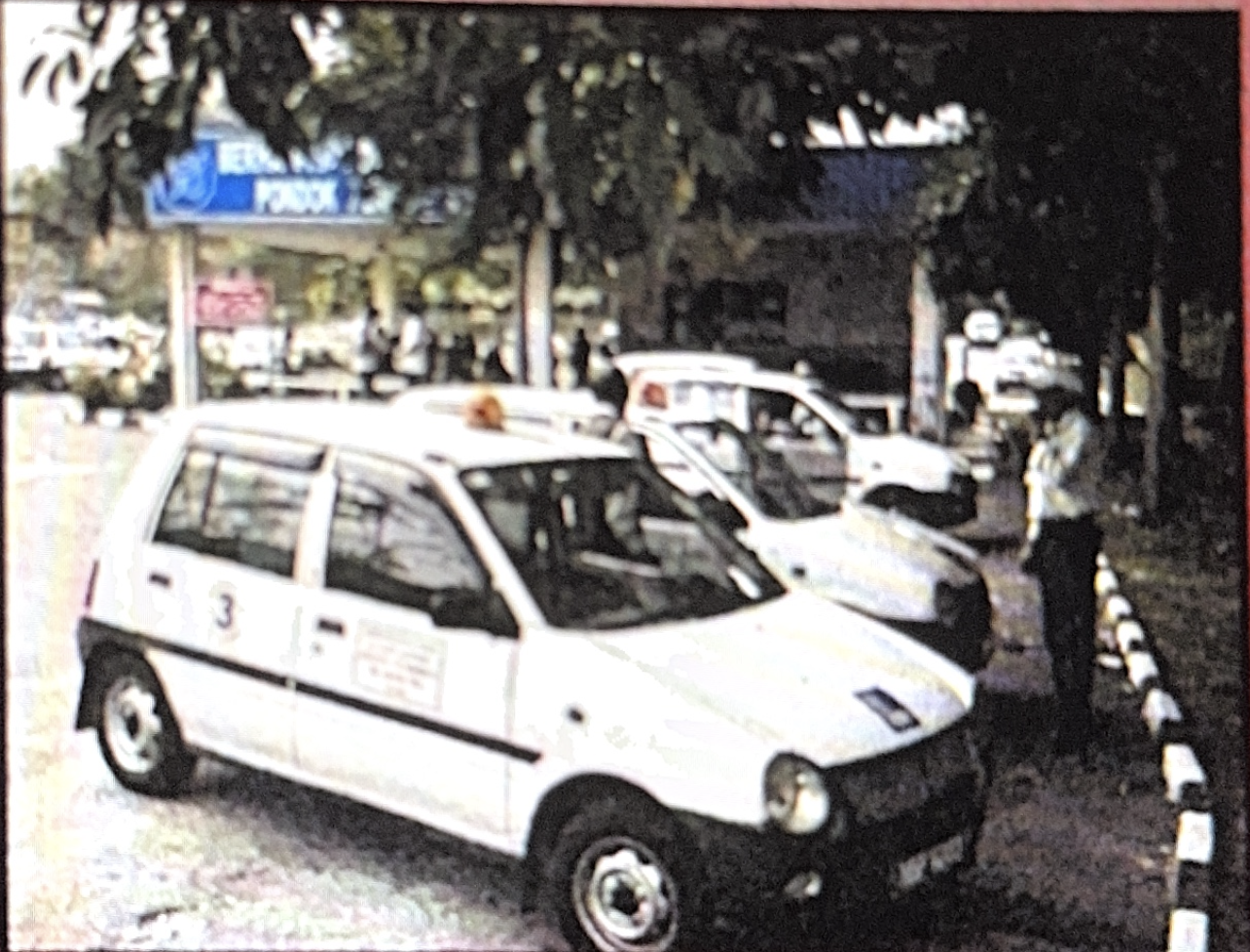 A parking lot with several white police cars parked in a row and people walking nearby, with trees and a blue sign in the background.