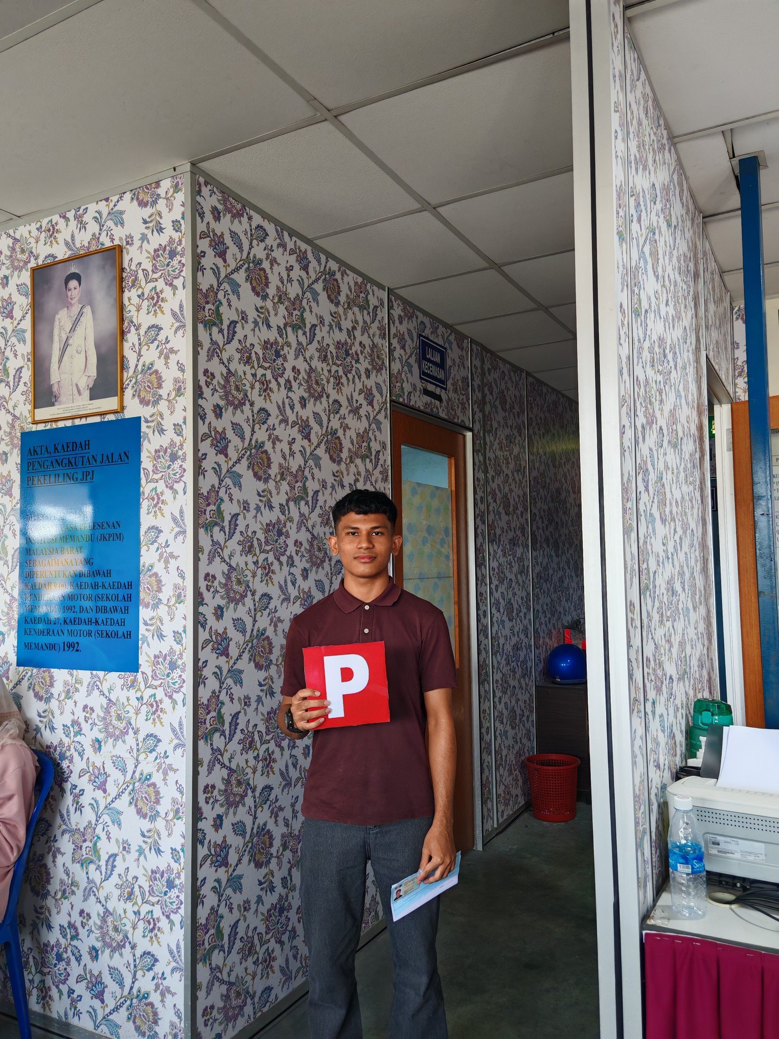 A young man standing inside a building, holding a red sign with a white letter P, next to a wall with floral wallpaper, and office furniture and equipment visible in the background.