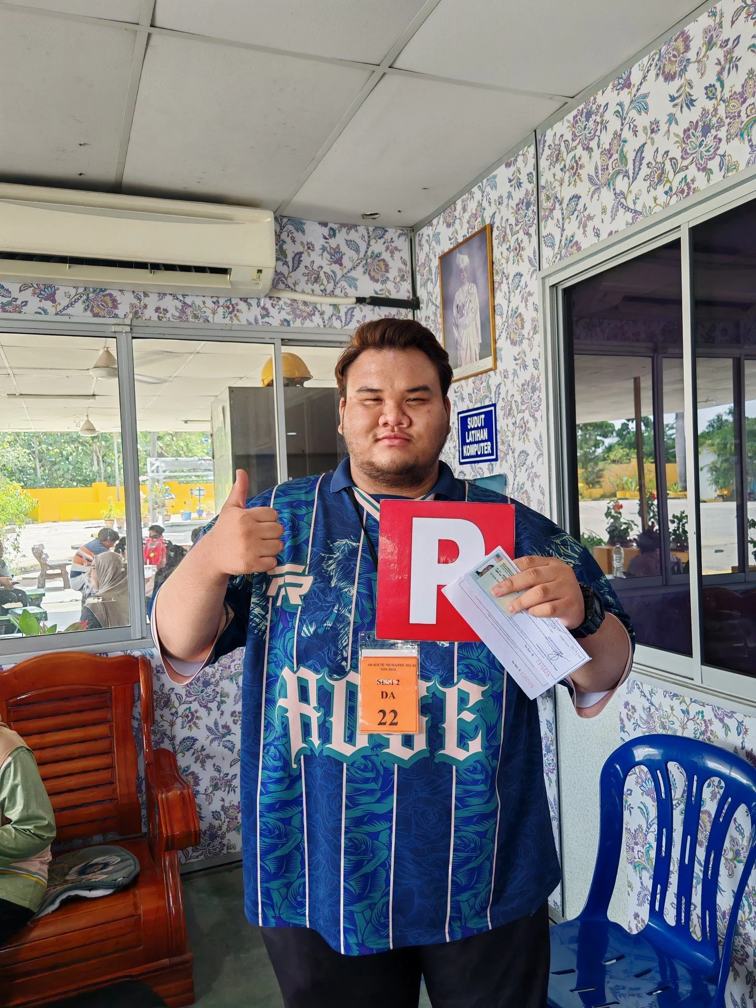 A young man holding a document and a red sign with the letter 'R' inside a building, giving a thumbs-up, with a floral patterned wall and glass windows behind him.