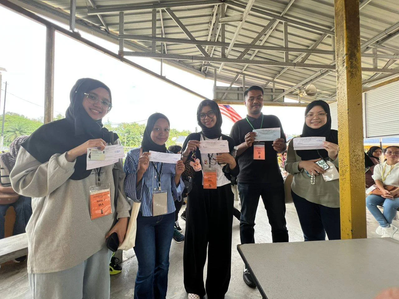 Five young adults, four women and one man, standing under a metal shelter, smiling and holding documents or tickets in their hands. They are wearing identification badges, and some women are wearing headscarves. Other people can be seen seated in the