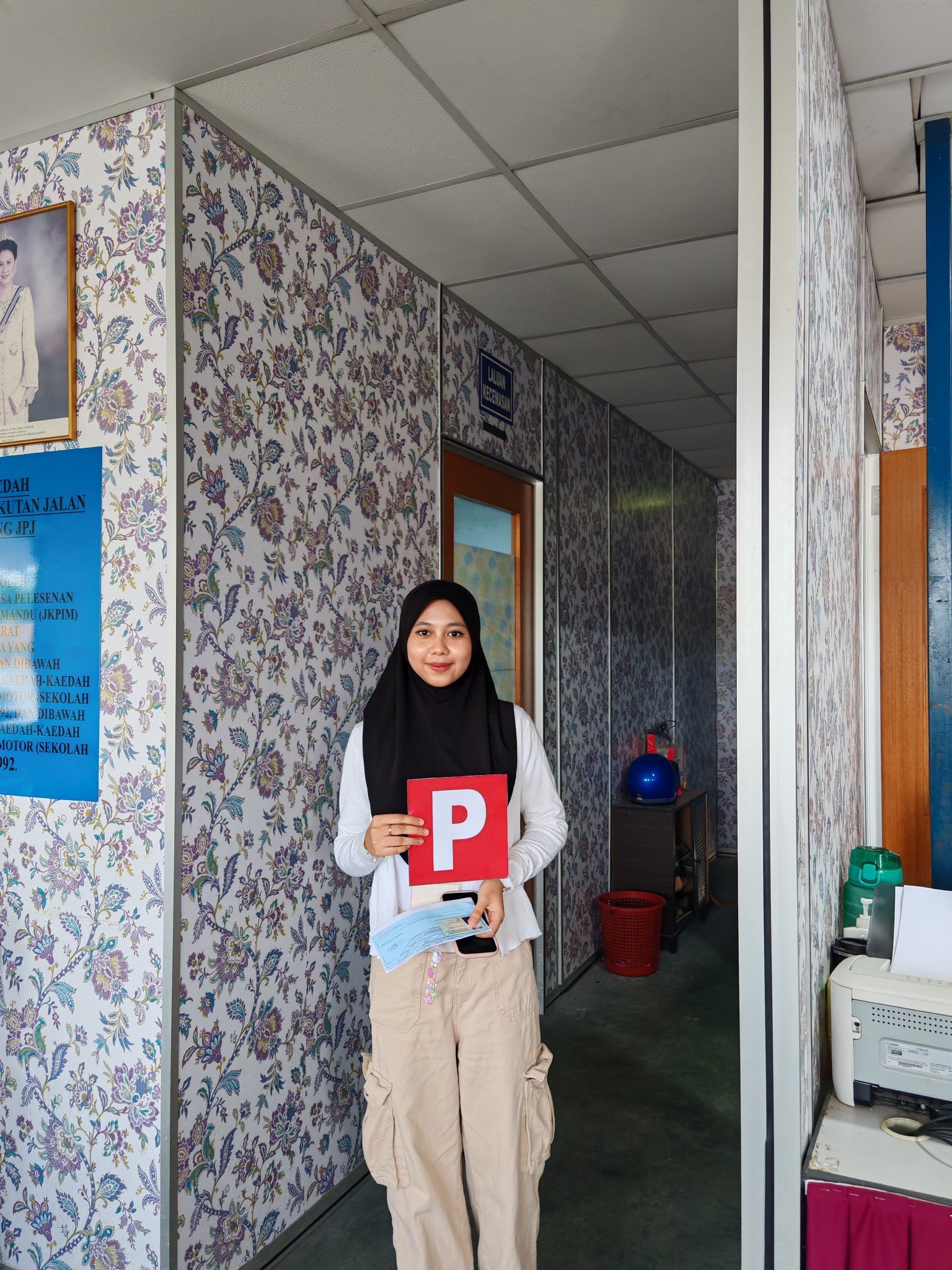 A woman standing indoors holding a red P sign and a piece of paper, wearing a black hijab and beige pants, in a room with floral wallpaper and office equipment.