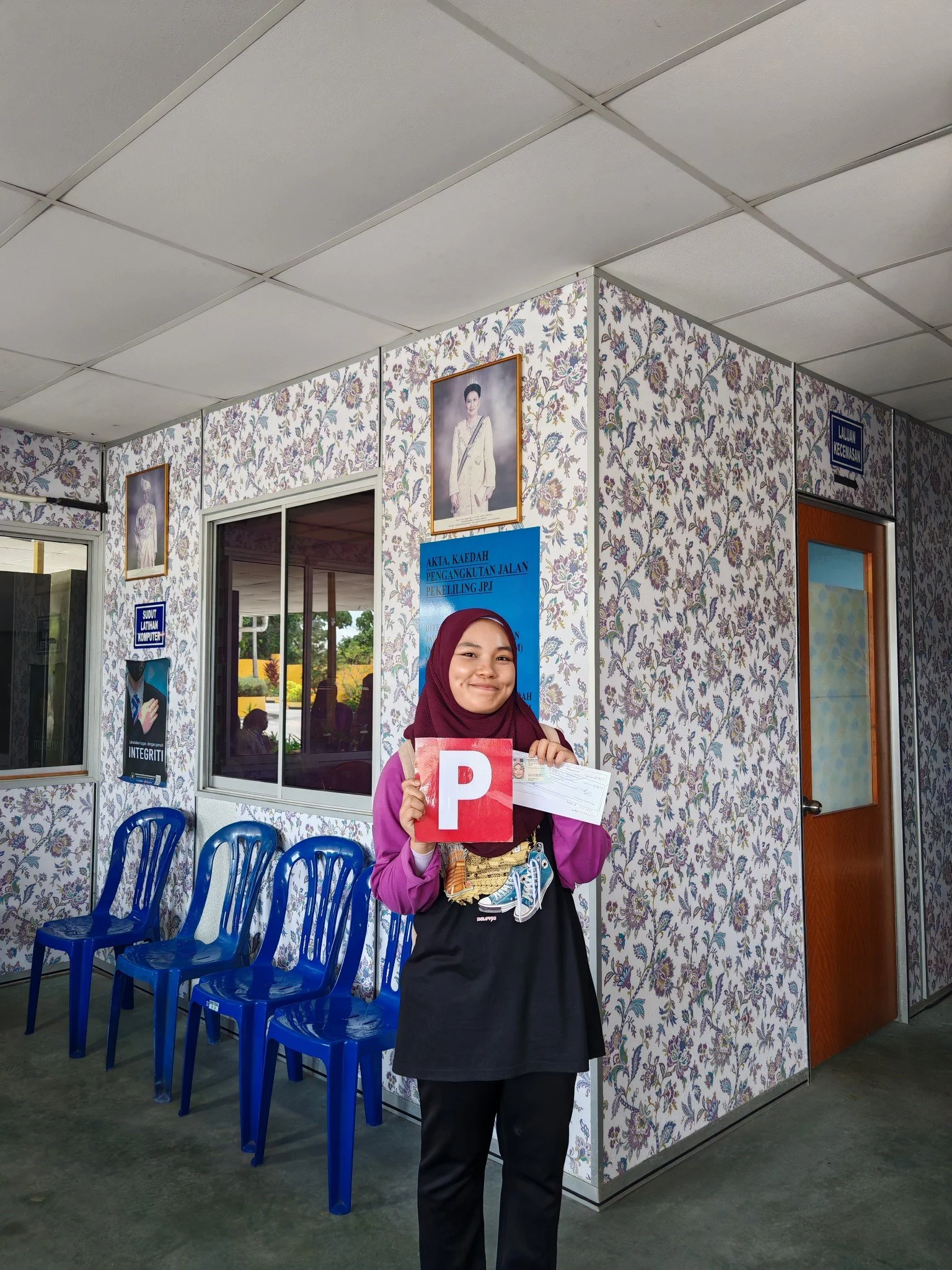 A young woman wearing a hijab and office uniform holding a pink 'P' sign and a check at a voting station.