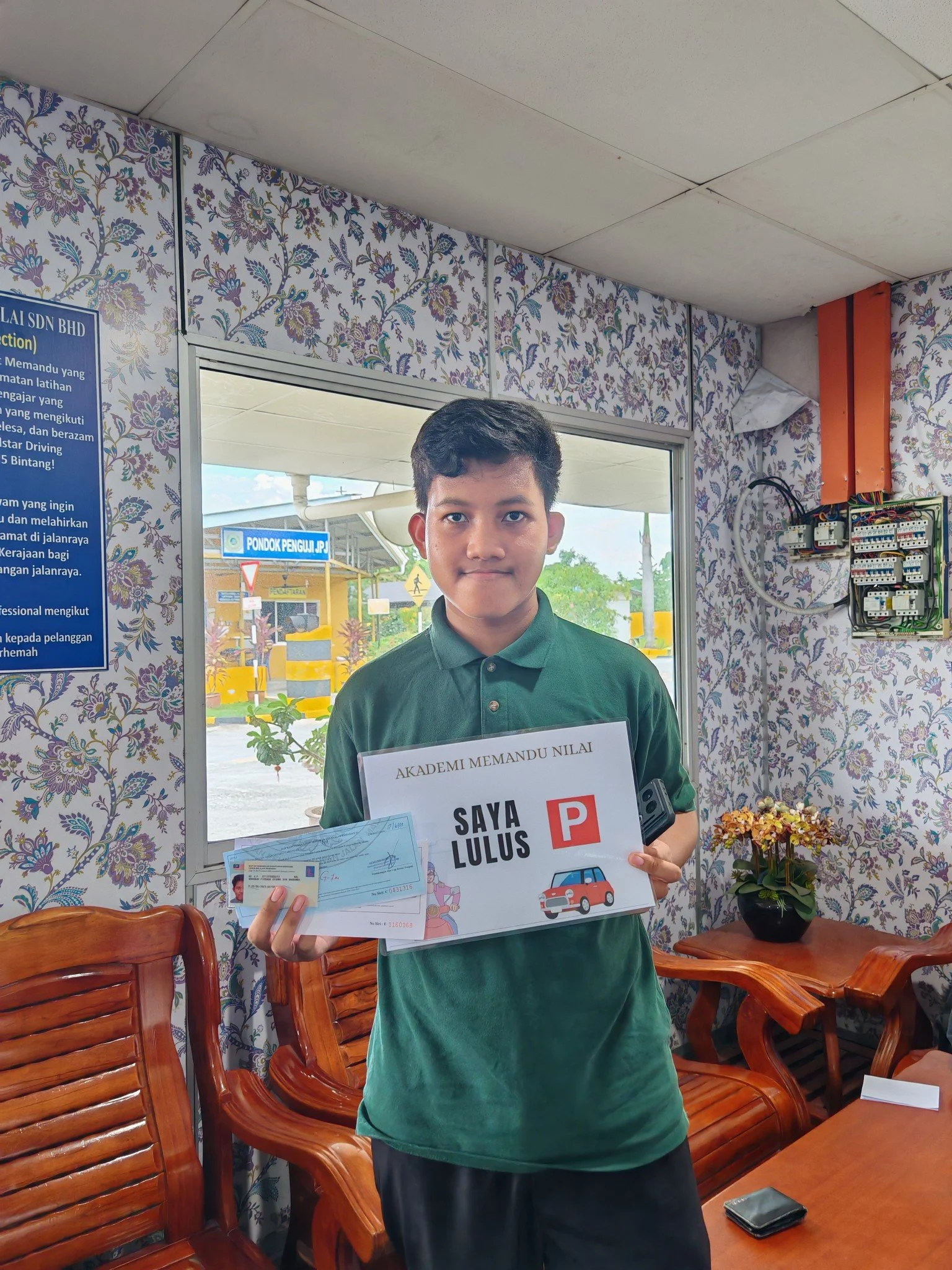 A young boy holding a driving test pass certificate, driver’s license, and a booklet in a room with floral wallpaper and wooden furniture.