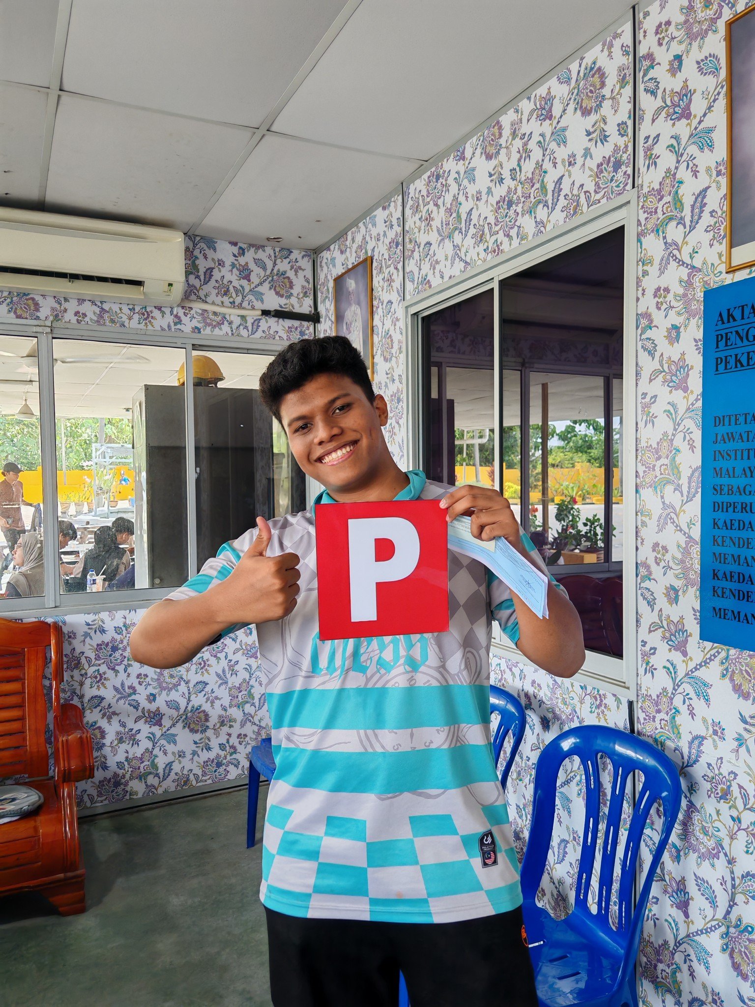 A young man smiling and giving a thumbs-up while holding a red and white sign with the letter 'P' in front of a patterned wall and blue chairs in a room.